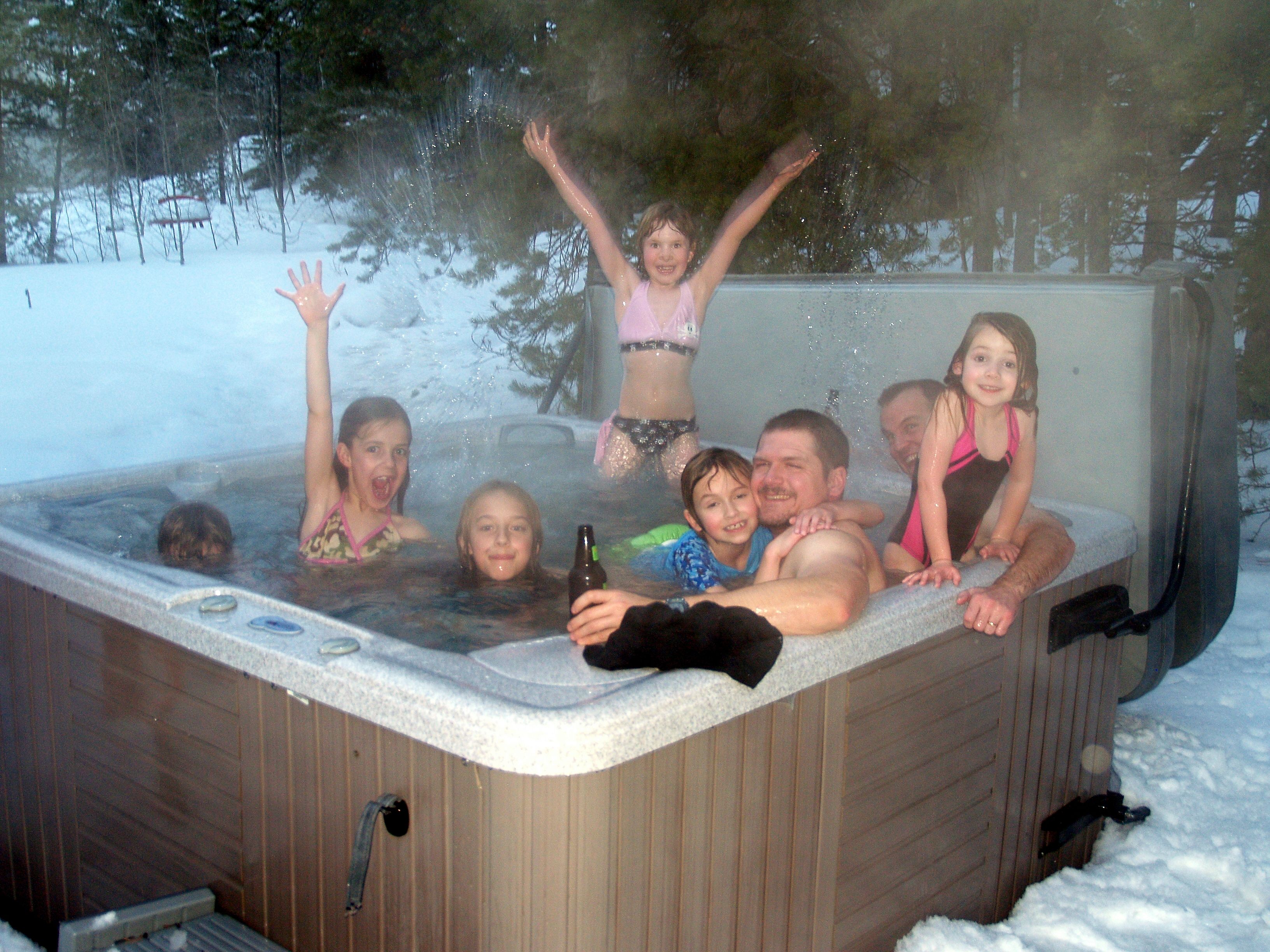 A group of seven people of mixed ages, children and adults, enjoying an outdoor hot tub in winter snow, with steam rising from the water and snow-covered conifers in the background; two of them have their arms raised in joy.