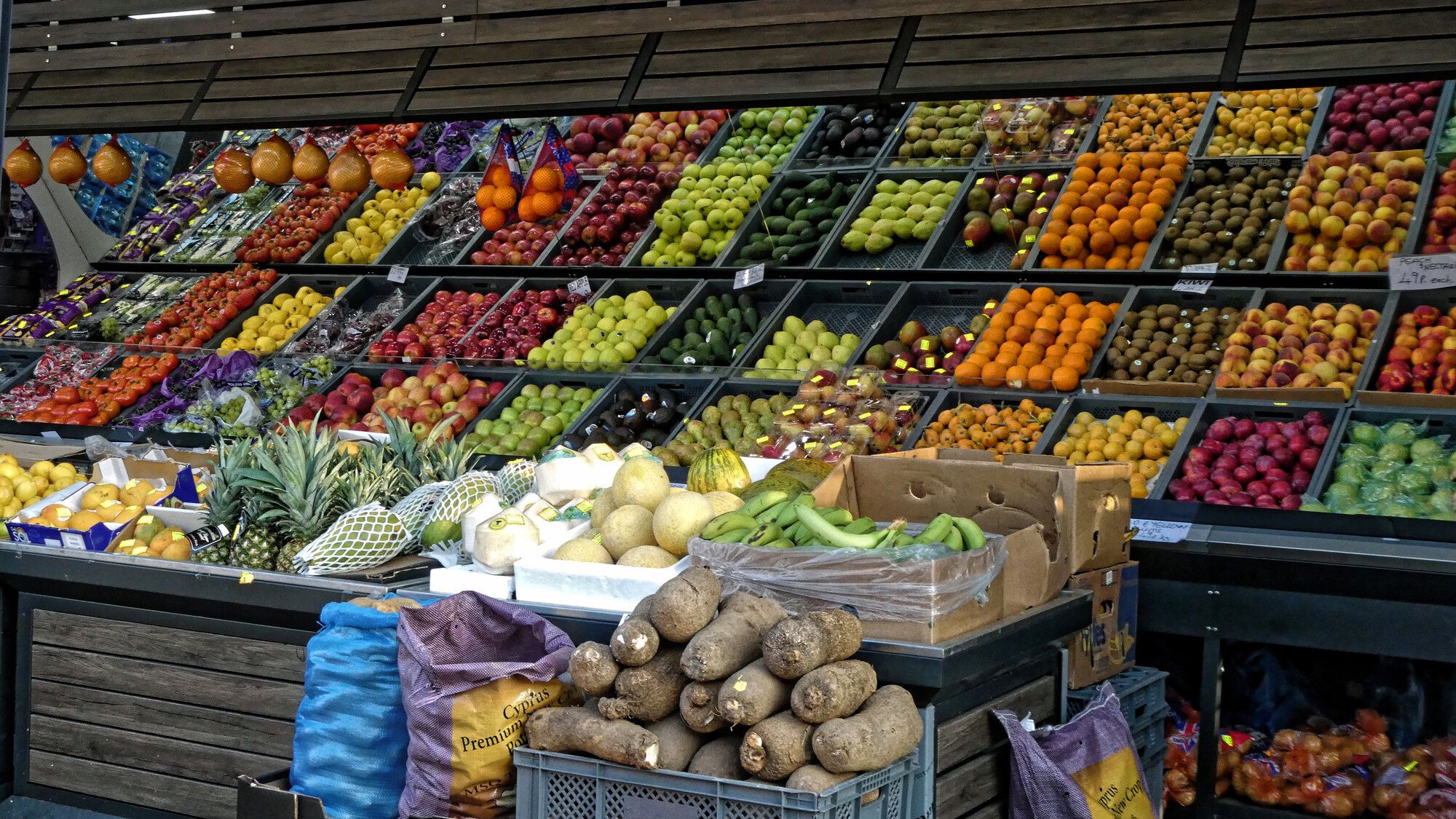 A greengrocer's display of crates of colourful fruit and vegetables stacked in rows, with sacks of root vegetables in the foreground.