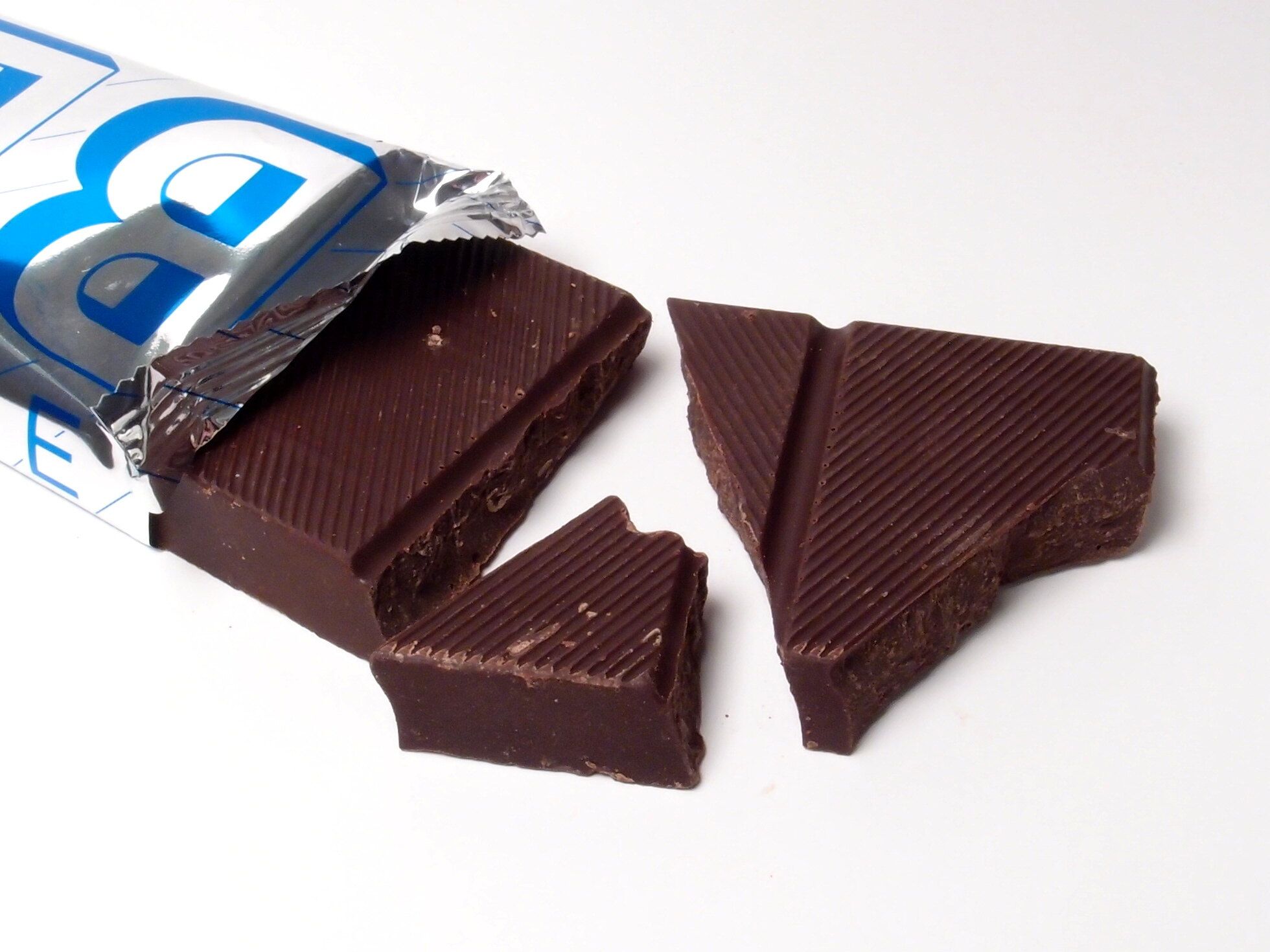 A dark chocolate bar in a half-opened blue and white foil wrapper, broken into pieces on a white surface.