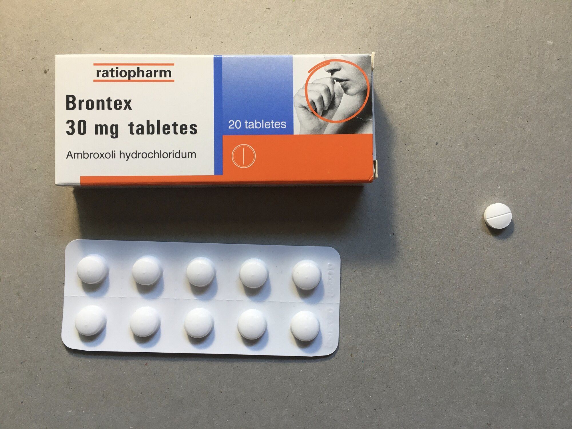 Κουτί φαρμάκου ratiopharm με μια κυψέλη (blister) από λευκά δισκία και ένα μεμονωμένο δισκίο, πάνω σε γκρι επιφάνεια.