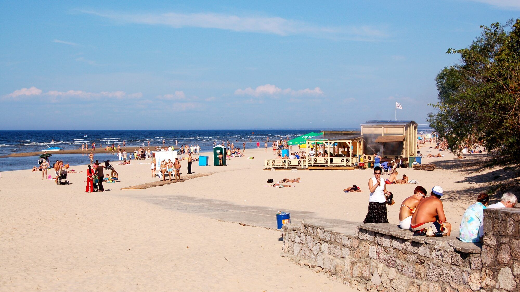 A sunny Baltic beach on a clear summer day, bathers scattered across pale sand and a calm sea meeting a blue sky, a wooden refreshment kiosk to the right.