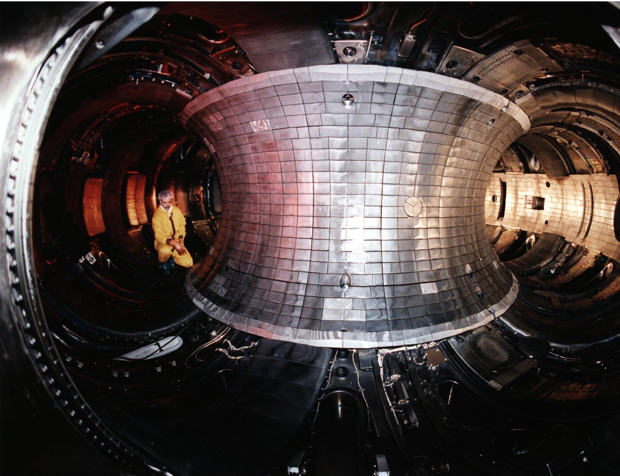 The mirror-bright, metal-tiled interior of a tokamak fusion reactor's doughnut-shaped vacuum vessel, with a technician in a yellow jacket crouched inside for scale.