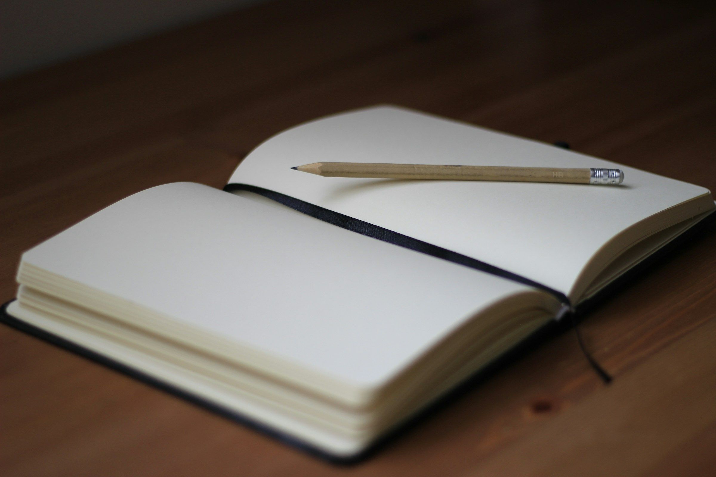 Open blank journal on a wooden table with a wooden pencil resting on the right-hand page and a thin black ribbon page-marker stretching across the gutter.