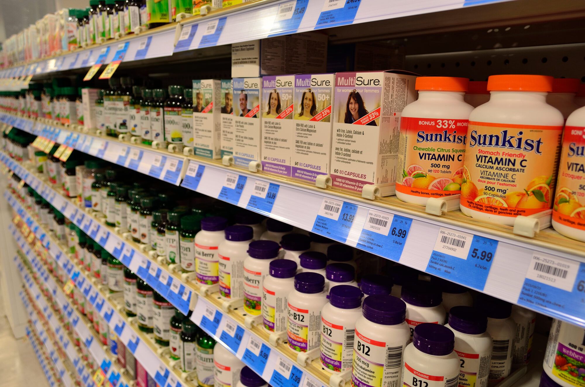 Pharmacy shelves packed with rows of vitamin and supplement bottles and boxes under bright store lighting, price tags along each shelf edge.